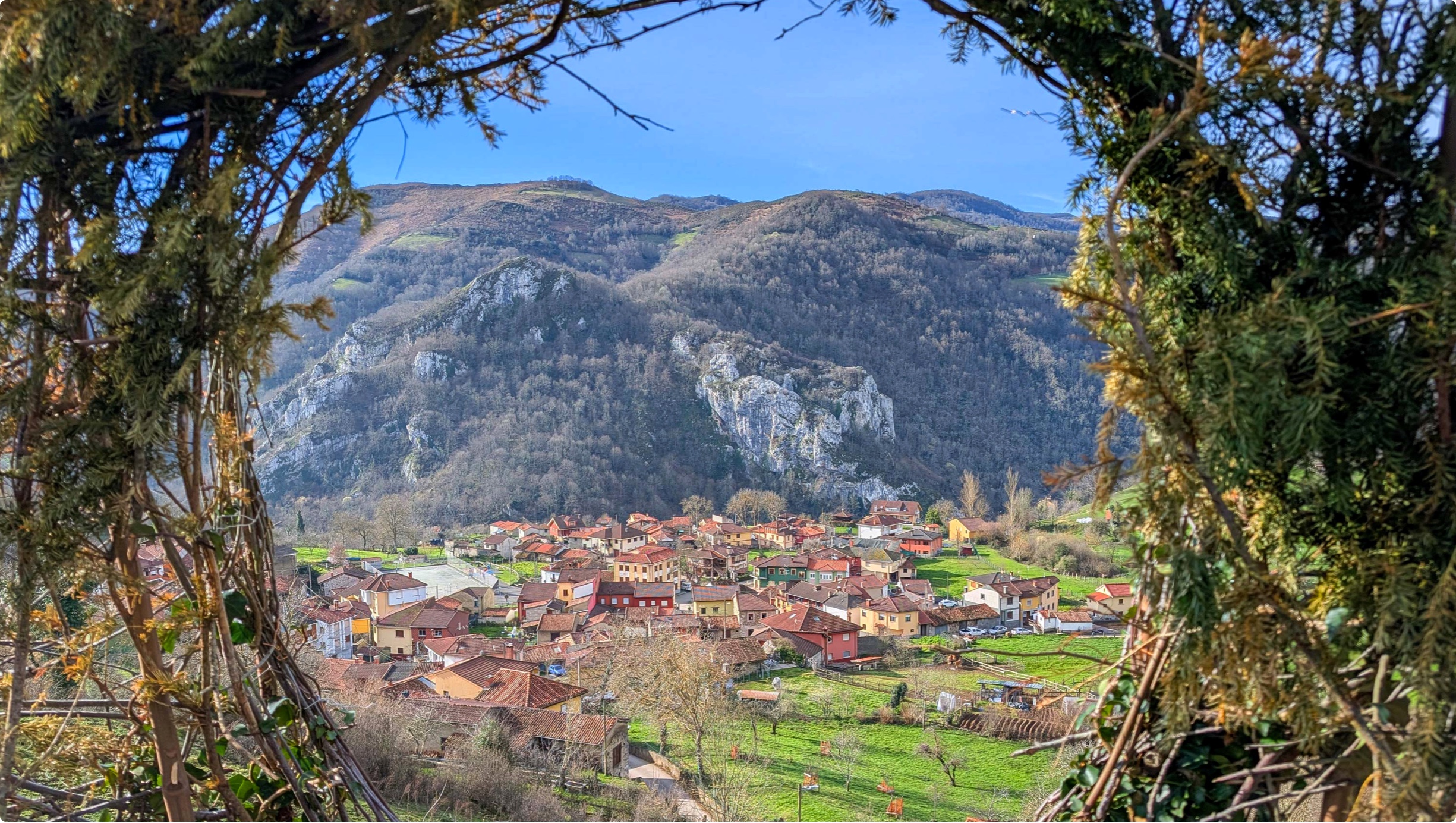 Vista panorámica de un pueblo rodeado de montañas y vegetación, enmarcada por ramas verdes