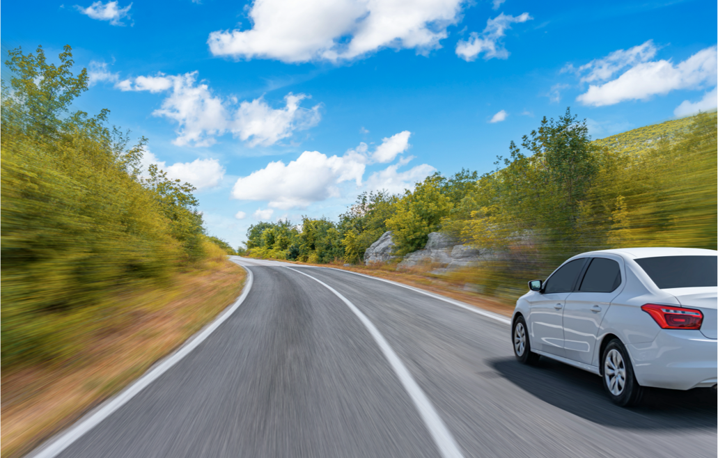 Coche blanco en movimiento por una carretera rodeada de árboles y un cielo azul con nubes