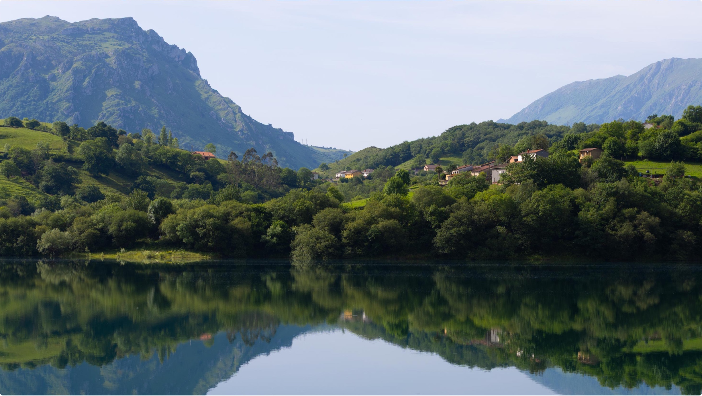 Paisaje montañoso con un lago reflejando colinas verdes y casas en un día soleado
