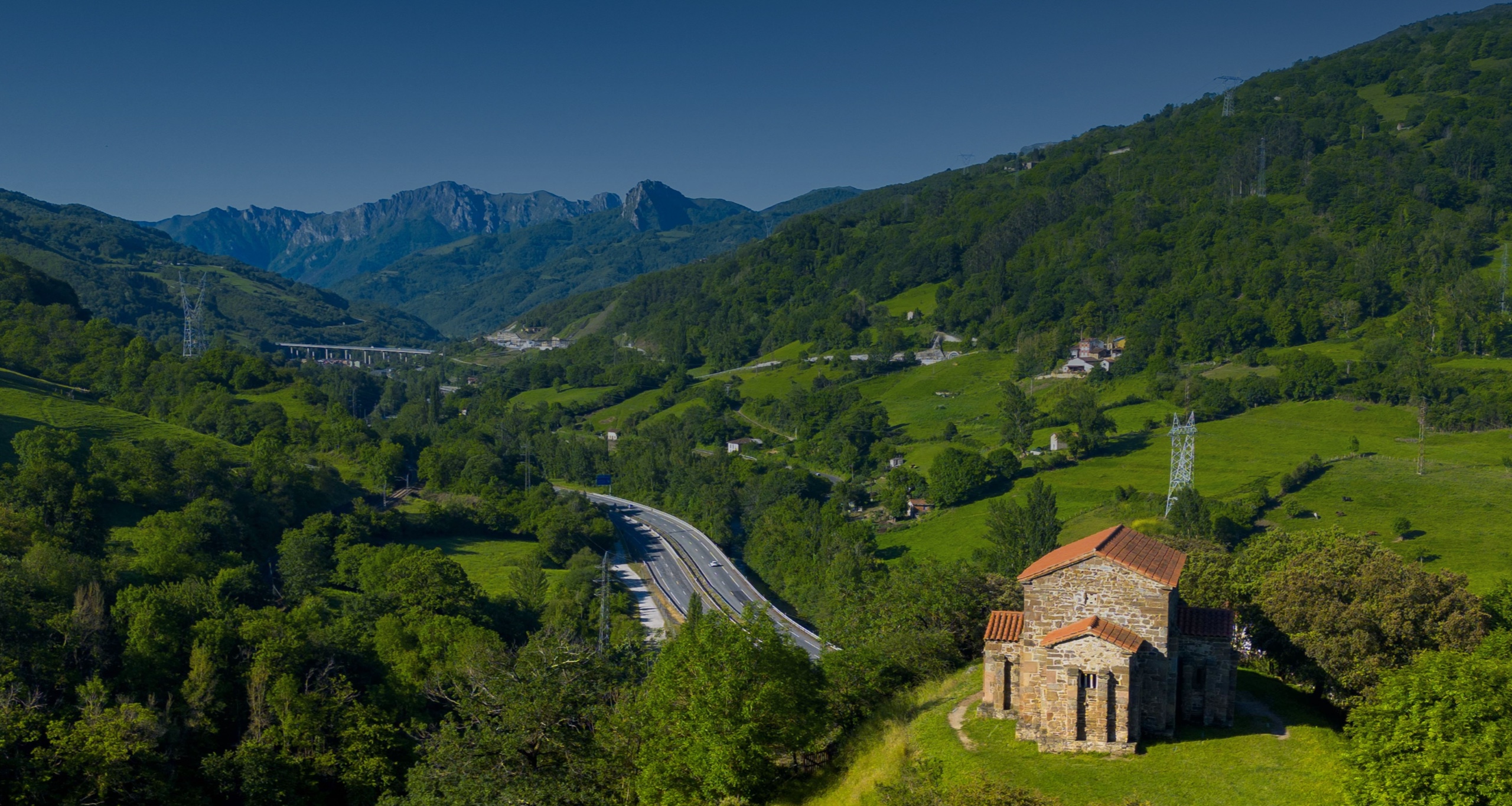 Iglesia de piedra en un paisaje montañoso con vegetación y una carretera en el fondo