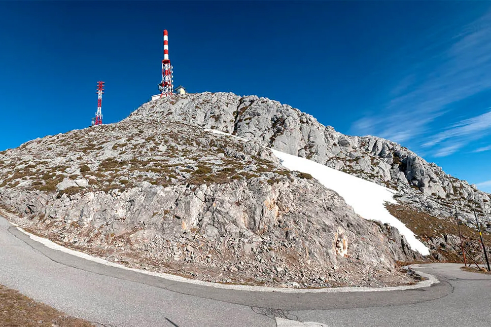 Montaña rocosa con torres de comunicación y un camino curvado en un día soleado