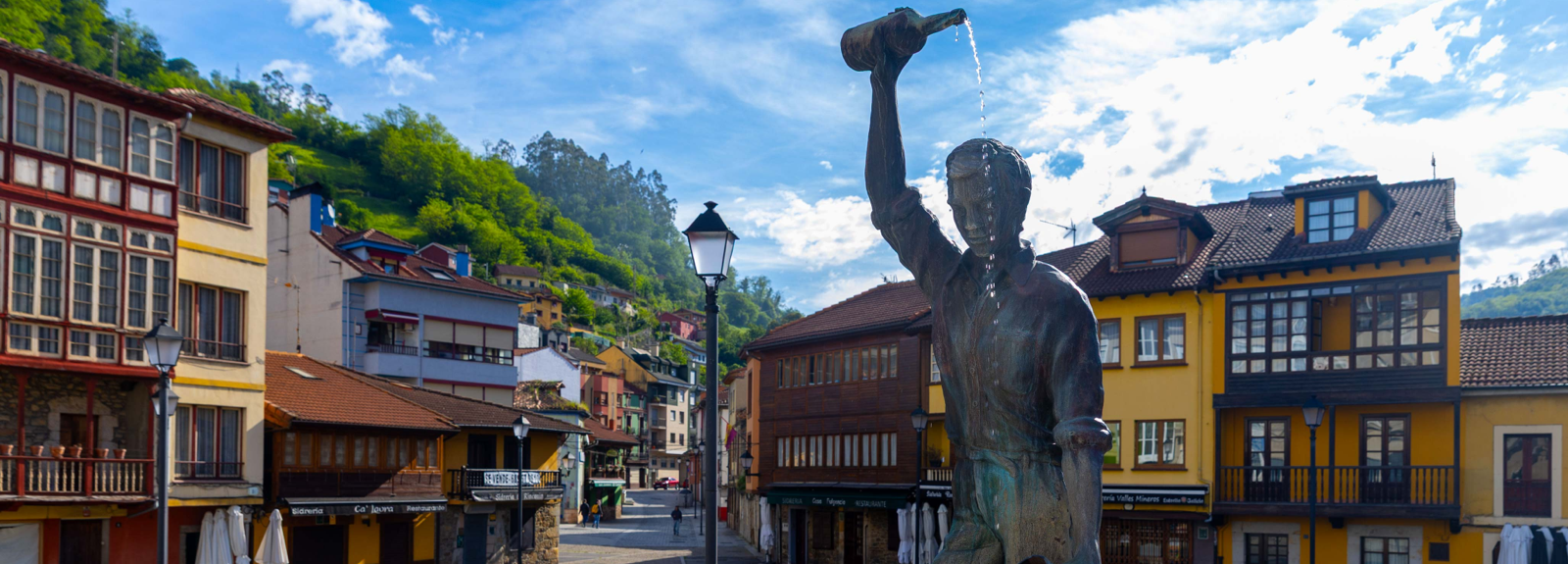 Estatua de un hombre sosteniendo una botella en una plaza rodeada de casas coloridas y montañas