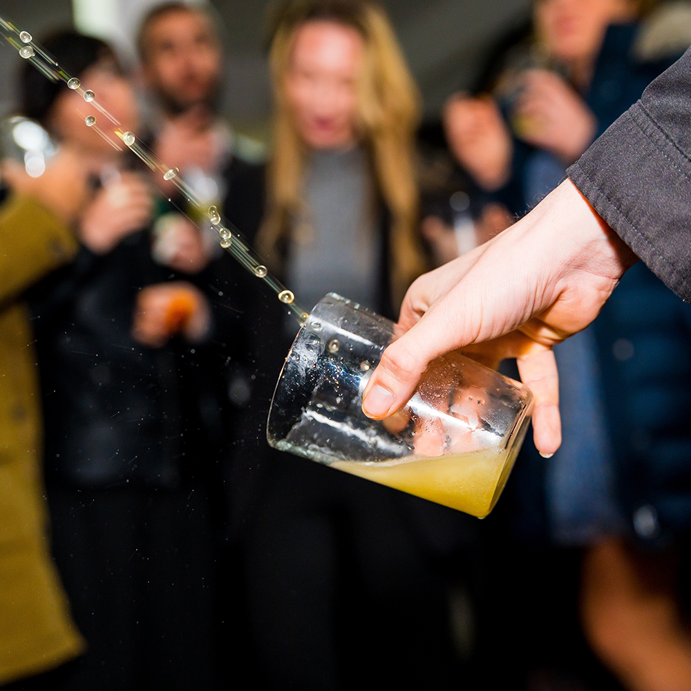 Vaso derramando bebida mientras un grupo de personas celebra de fondo