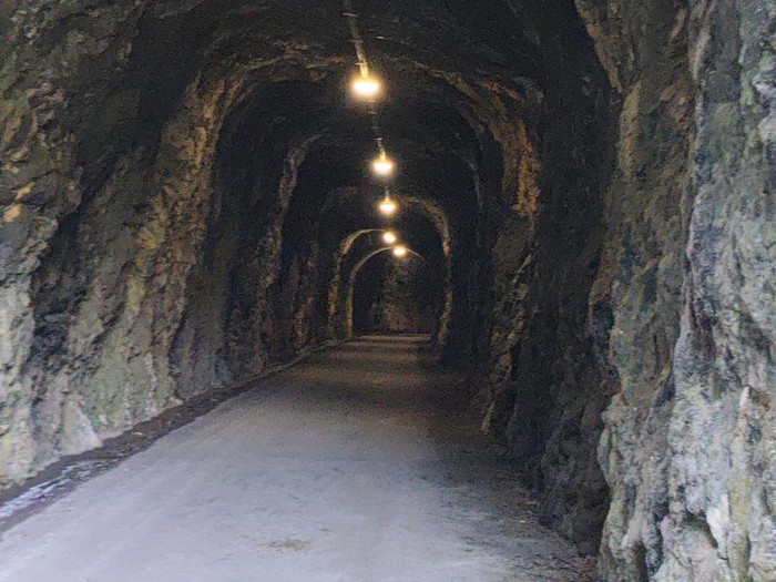 Túnel iluminado con luces en el techo y paredes de roca, con un camino de tierra en el centro