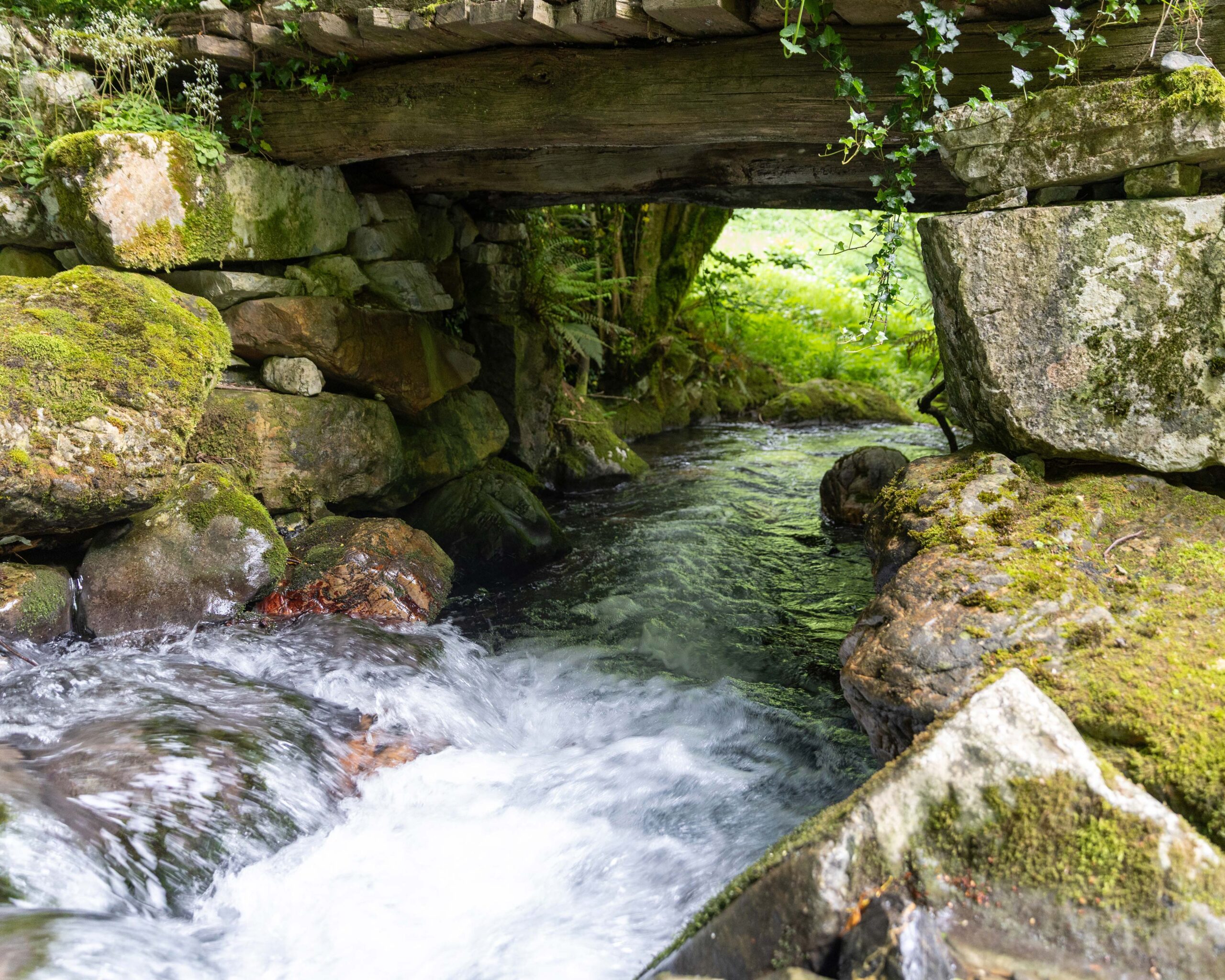Río fluyendo bajo un puente de madera, rodeado de rocas y vegetación