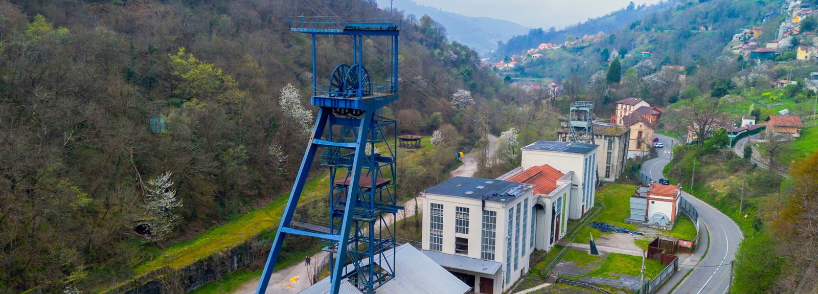 Estructura de un antiguo pozo minero rodeada de vegetación y edificios industriales en un paisaje montañoso