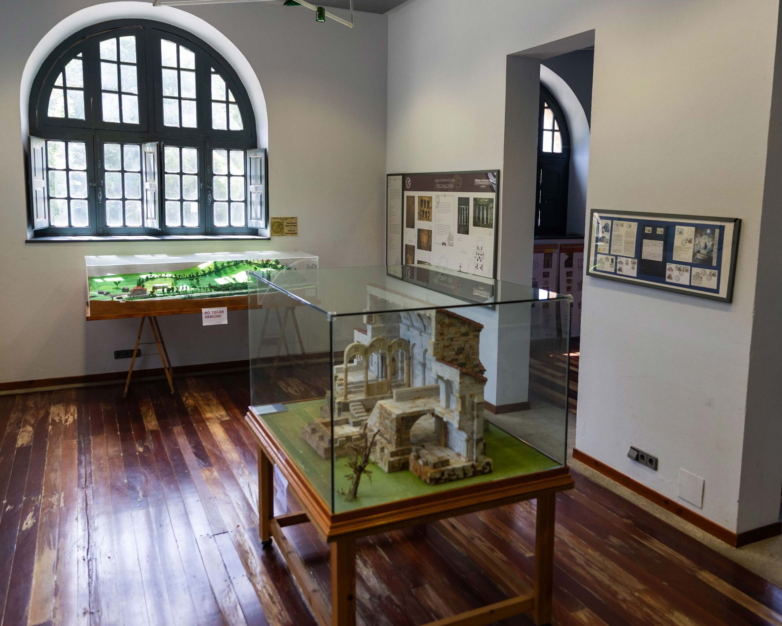 Sala de exposición con maquetas y paneles informativos, ventanas arqueadas y piso de madera