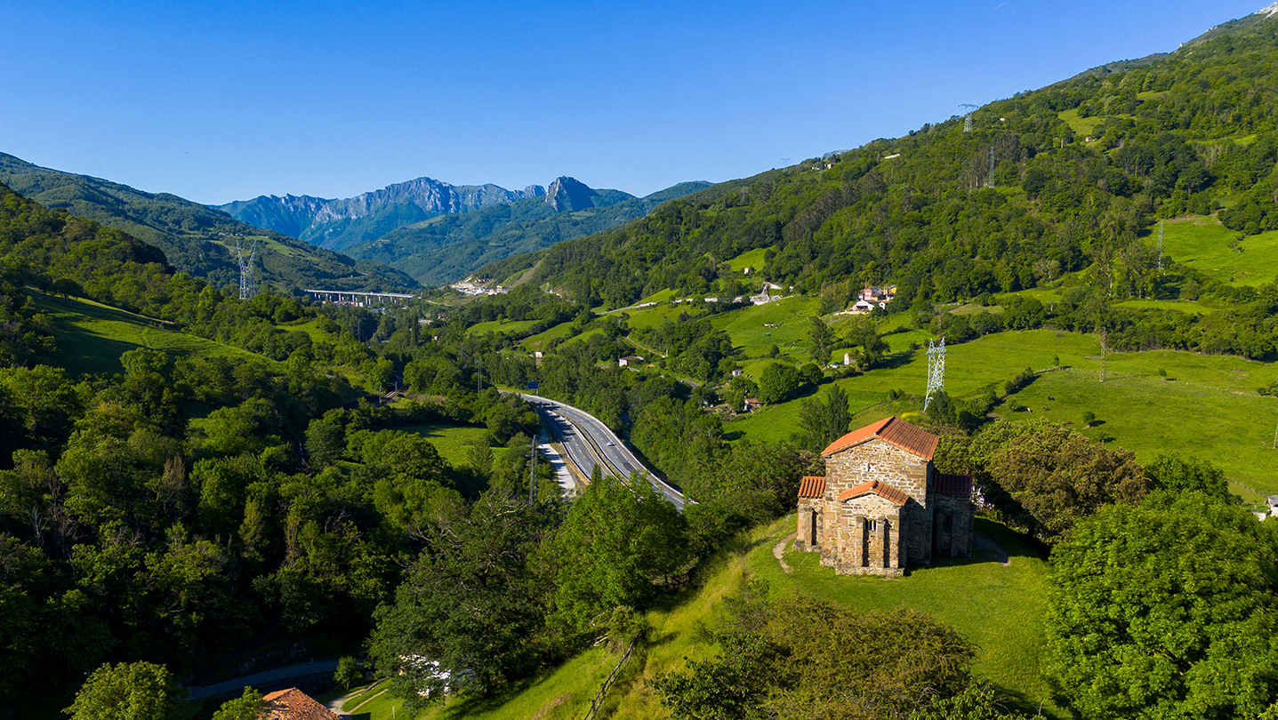 Iglesia de piedra en un paisaje montañoso verde con carretera y montañas al fondo