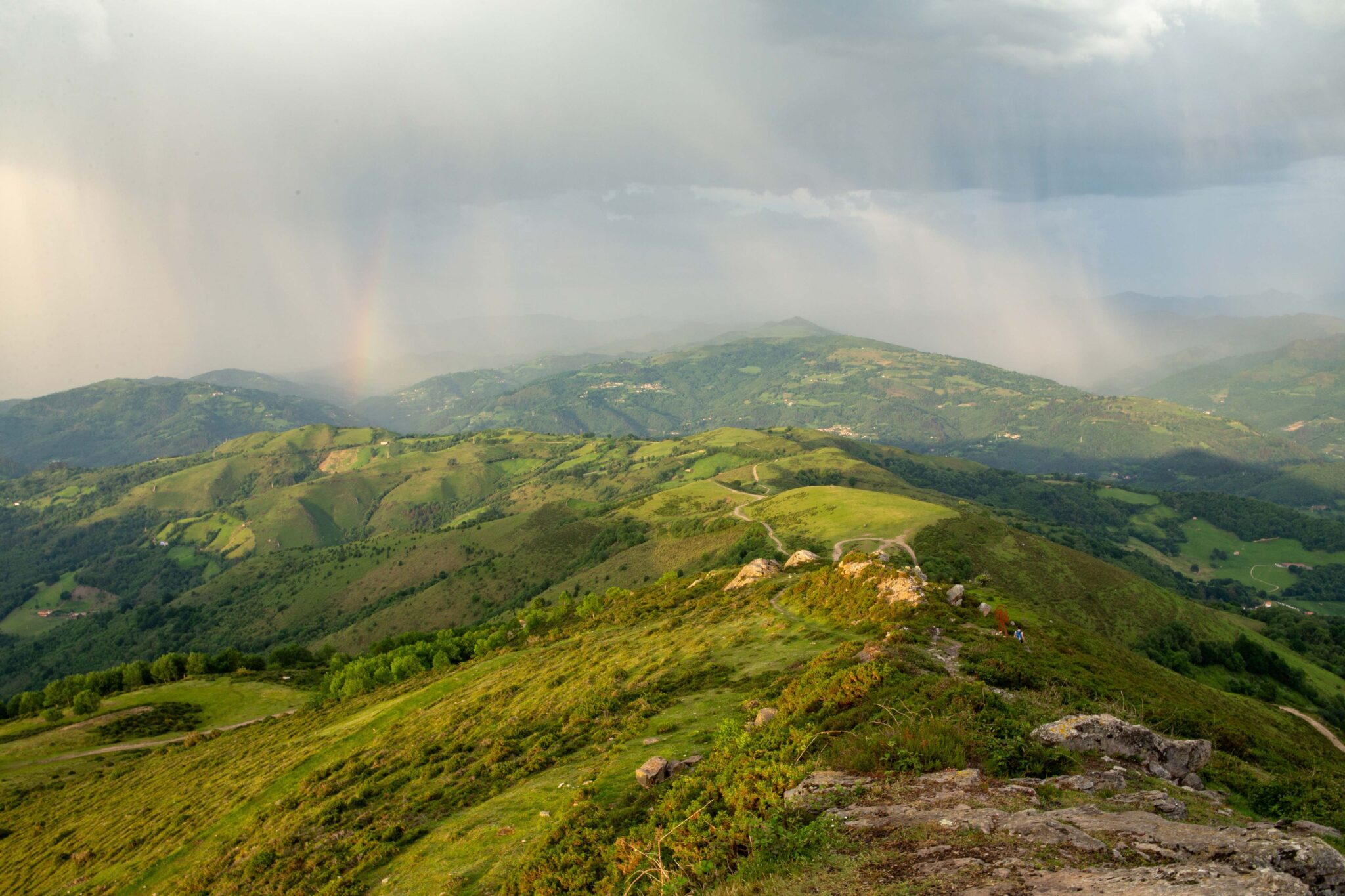 Paisaje montañoso con colinas verdes, cielo nublado y un arcoíris en la distancia