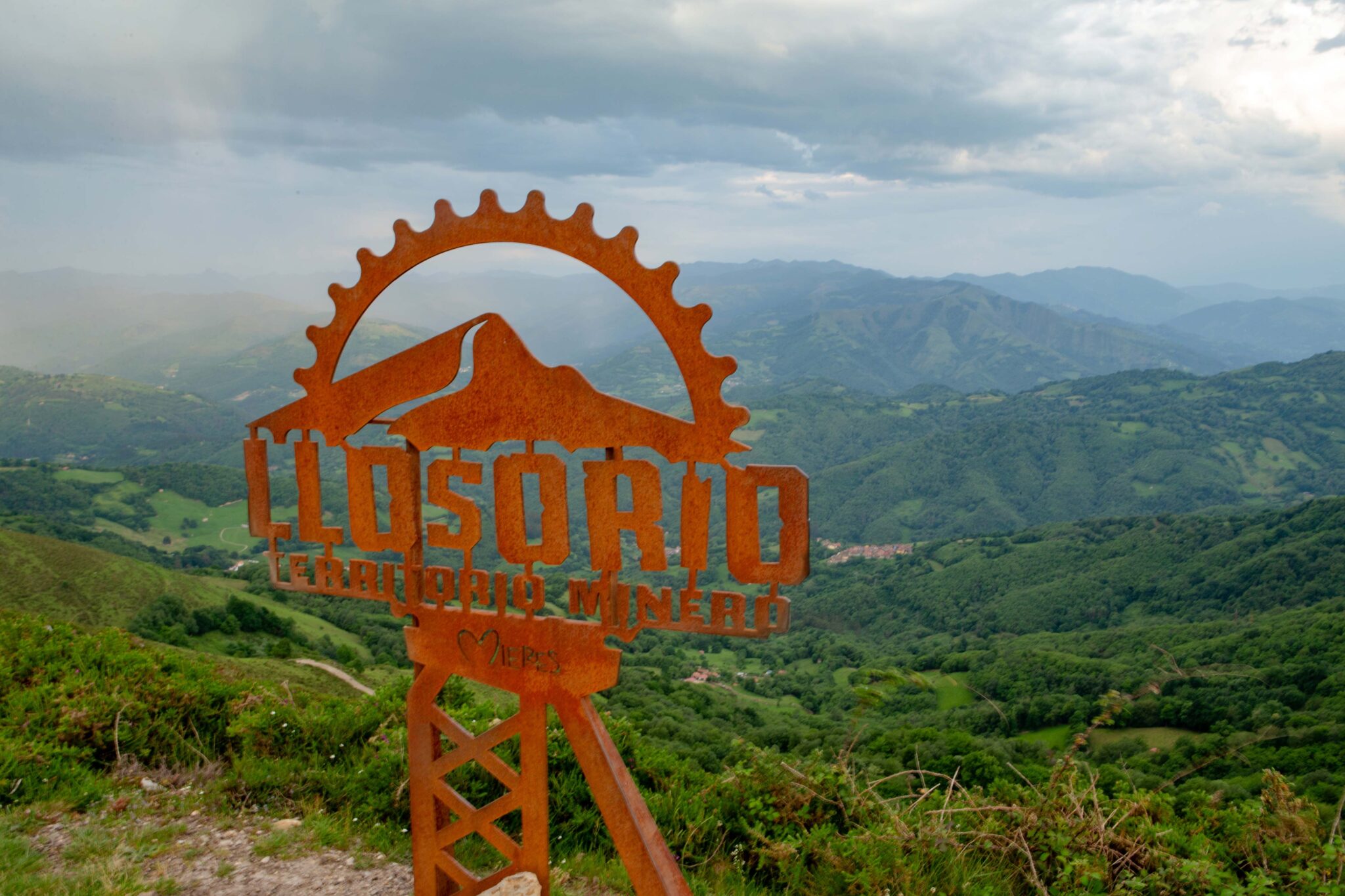Cartel de metal oxidado que dice "Llosorio" con un paisaje montañoso al fondo