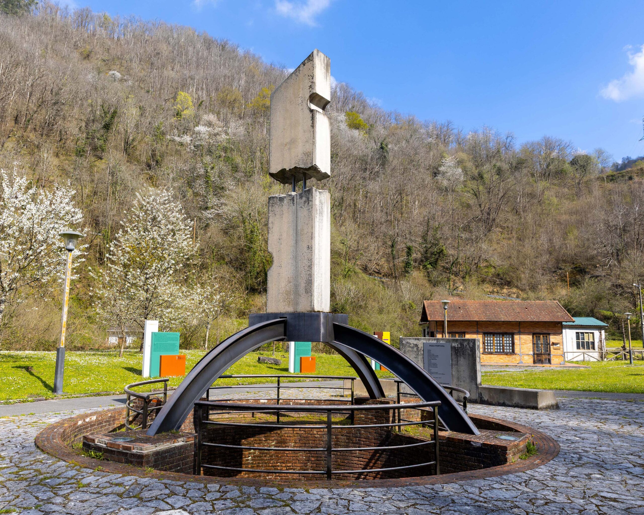 Monumento abstracto en un parque rodeado de vegetación y una cabaña de madera al fondo