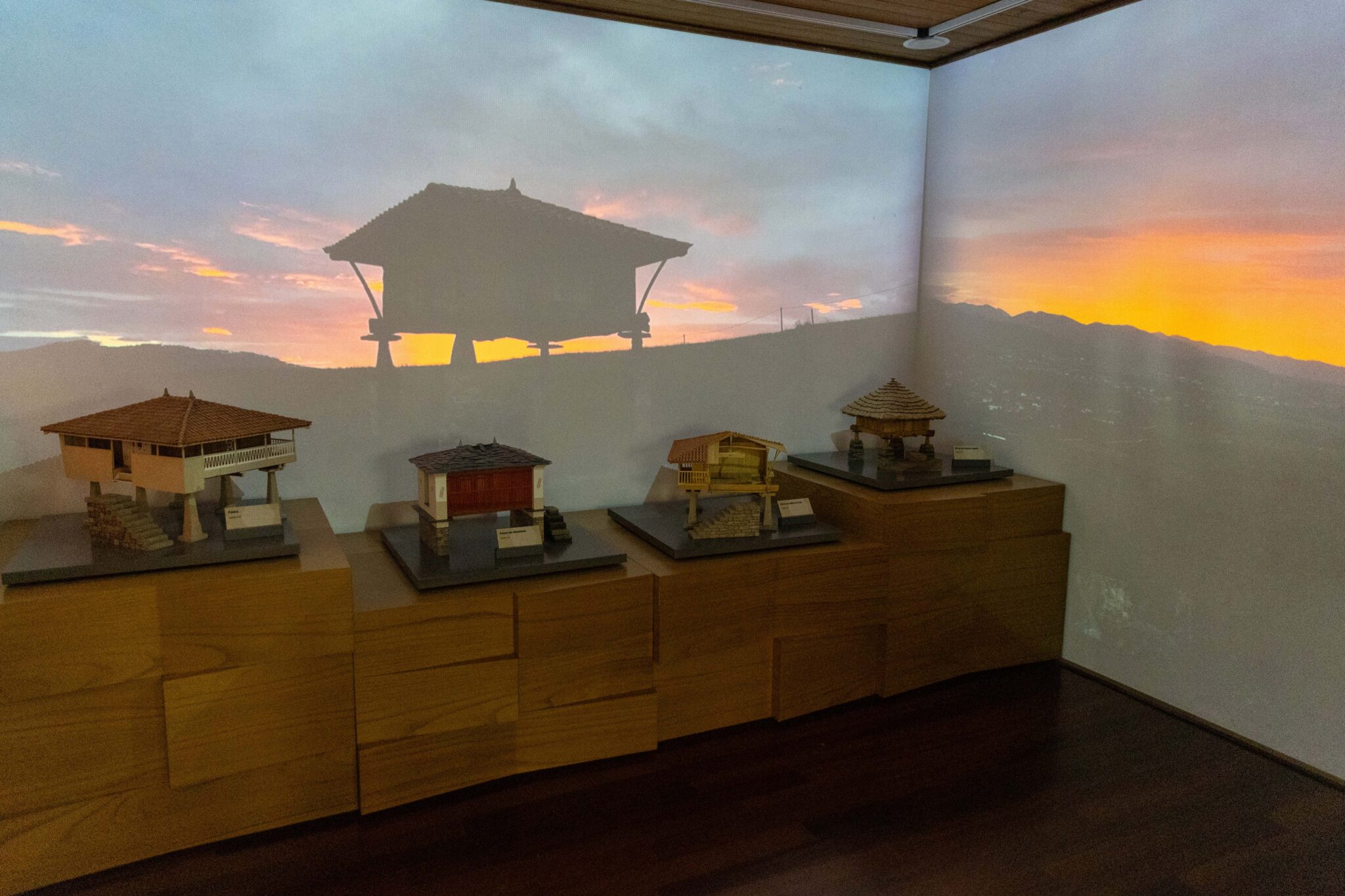 Maquetas de casas en una sala con proyección de un atardecer en el fondo