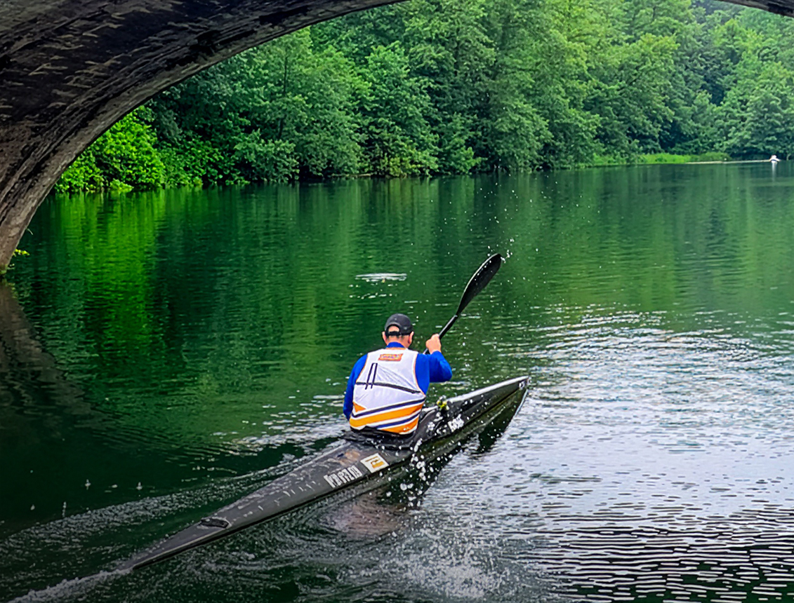 Hombre remando en kayak bajo un puente, rodeado de vegetación y agua tranquila