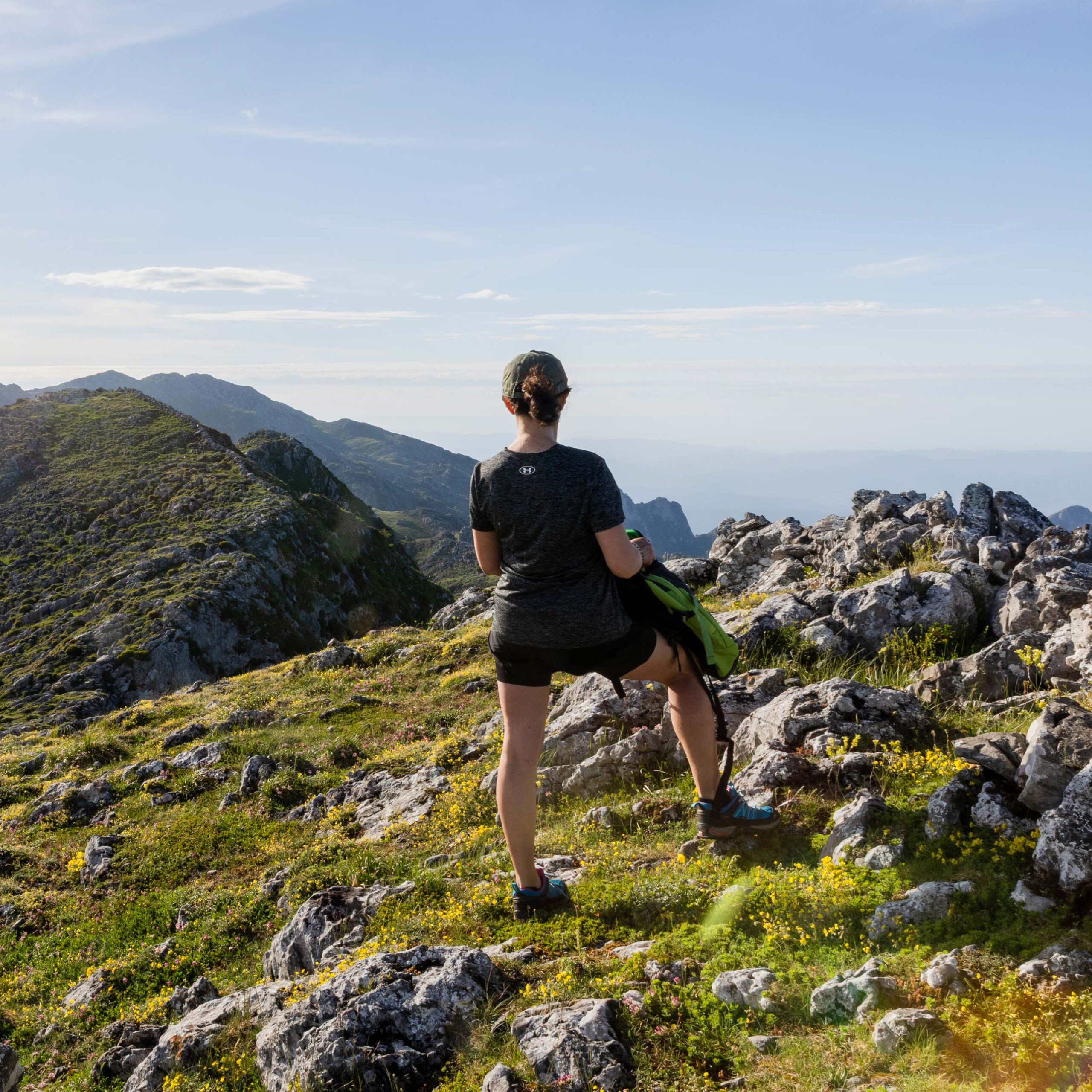 Persona de espaldas en una cima montañosa, rodeada de rocas y vegetación, contemplando el paisaje