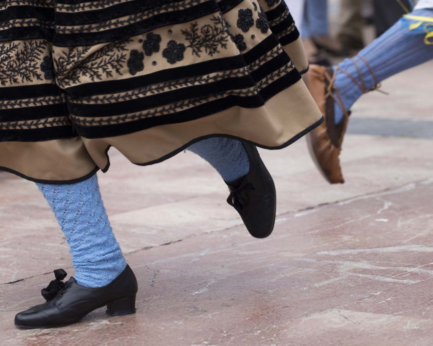 Piernas de personas con trajes tradicionales, una con falda negra y otra con pantalones y zapatos de cuero