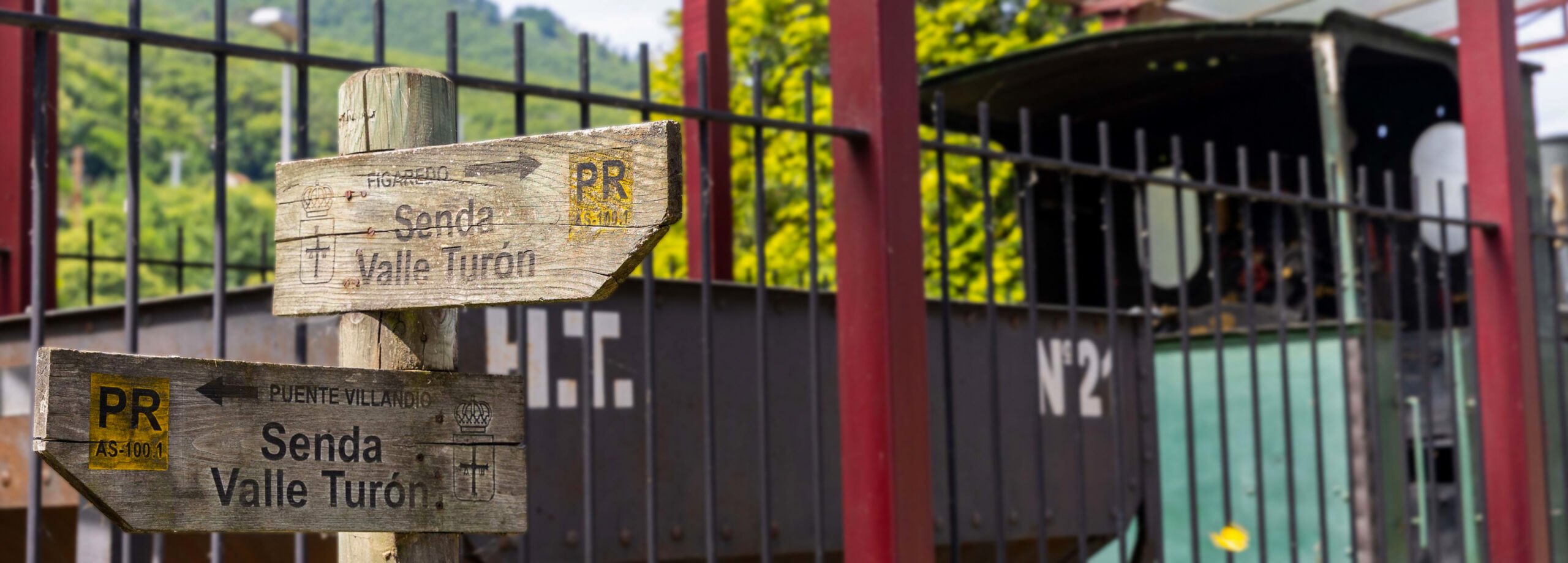 Señales de madera indicando direcciones hacia Valle Tirón y Senda Valle Turón, con un fondo de tren antiguo