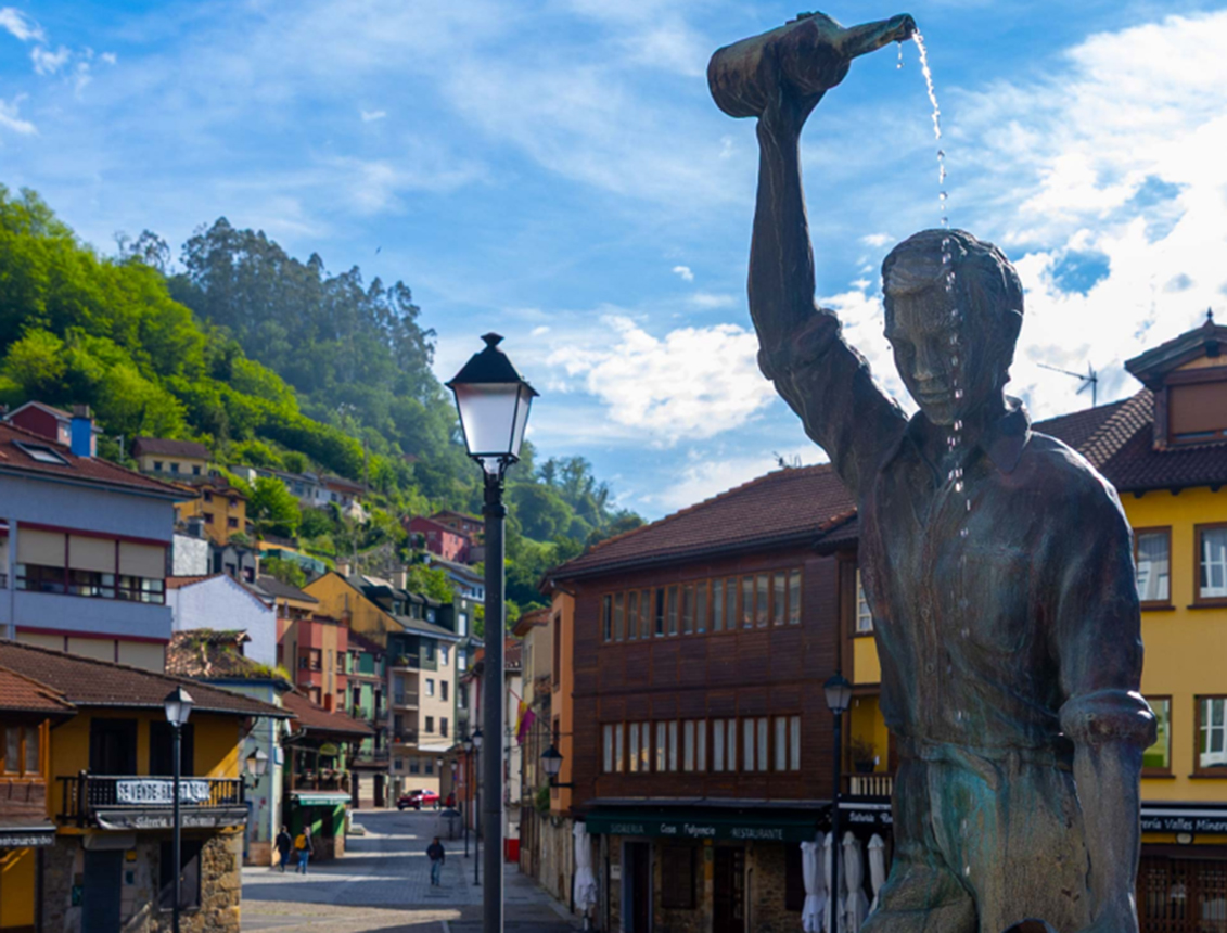 Estatua de un hombre vertiendo líquido en una plaza con casas coloridas y montañas al fondo