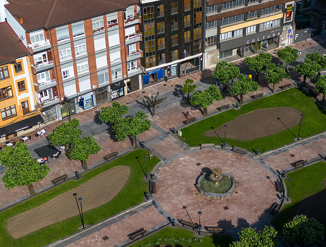 Plaza con árboles, bancos y una fuente, rodeada de edificios en un día soleado