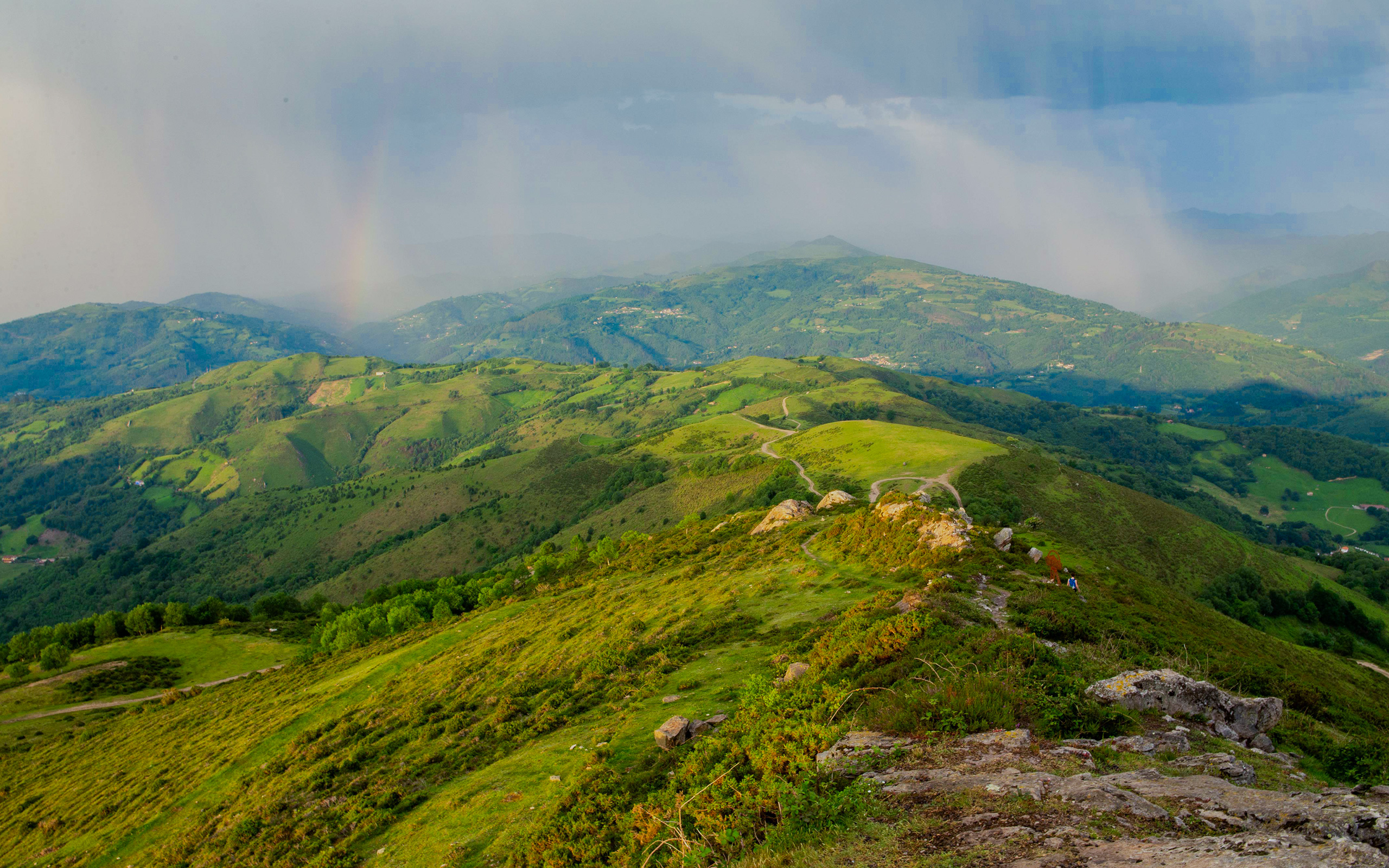 Vista panorámica de montañas verdes con un arcoíris en el horizonte y nubes en el cielo