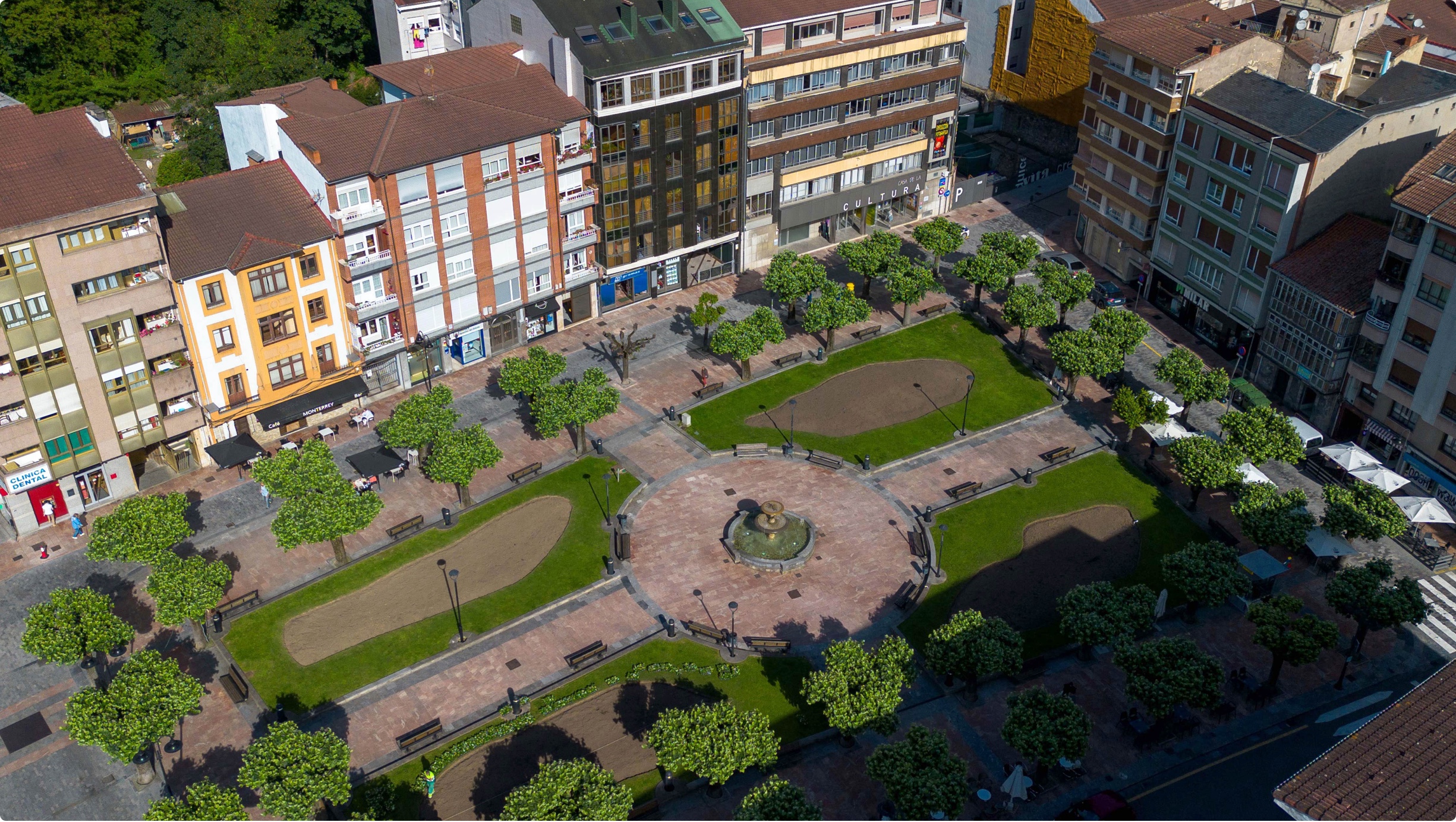 Plaza con árboles, bancos y una fuente, rodeada de edificios en un entorno urbano