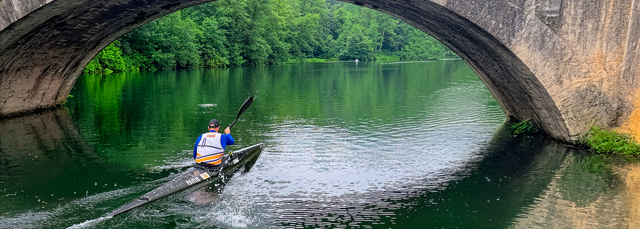 Remero en un kayak navegando por un río bajo un puente, rodeado de vegetación