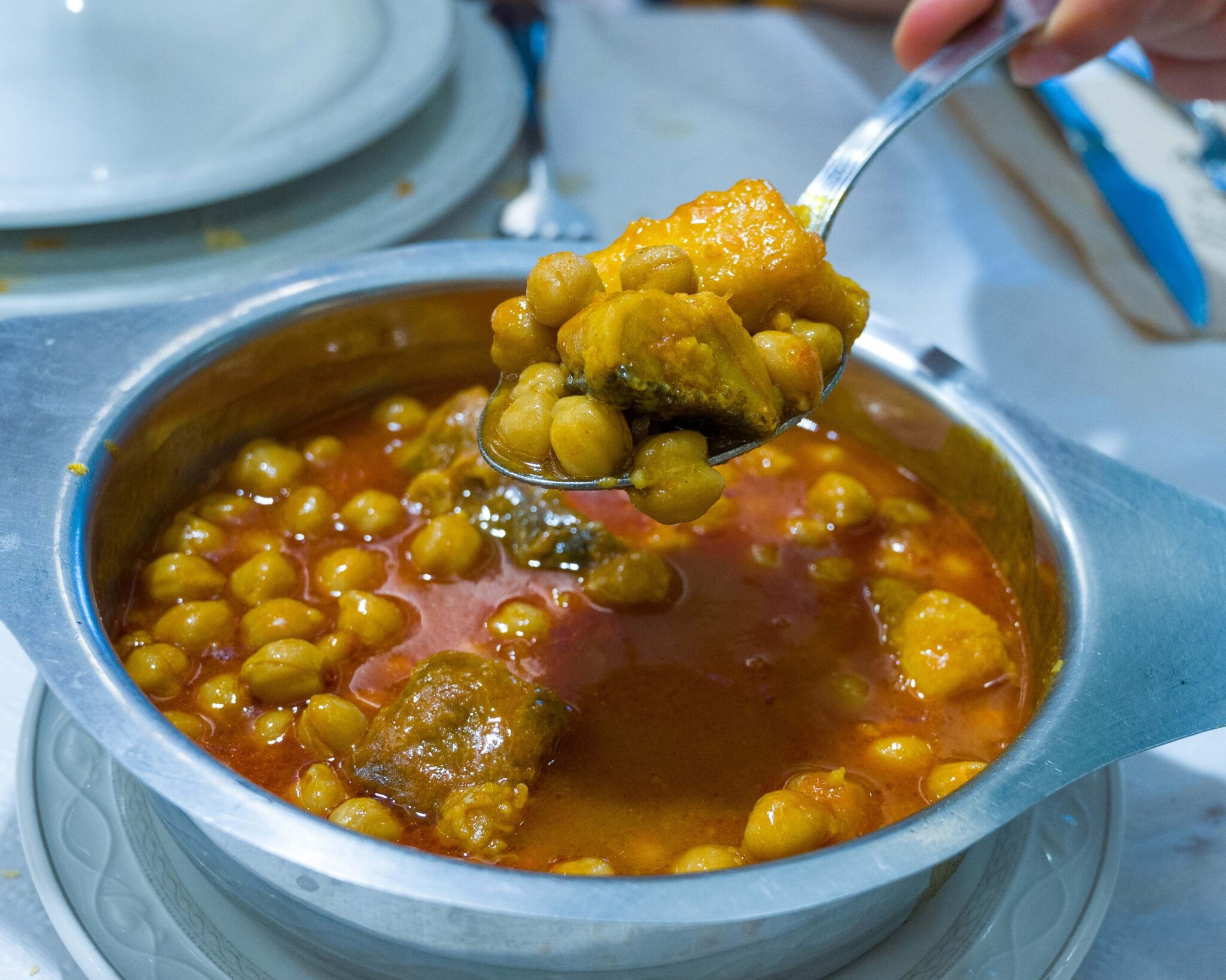 Sopa caliente con garbanzos y trozos de carne en un recipiente metálico, con una cuchara sirviendo
